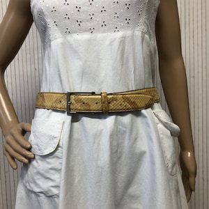 Peut inclure: Une robe blanche à œillets avec une ceinture en peau de serpent beige et une boucle argentée. La robe est sans manches et a une jupe évasée. La ceinture est portée à la taille, soulignant la silhouette de la robe. La robe a des poches.