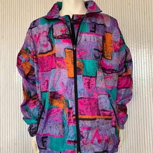 Puede incluir: Chaqueta vintage con cremallera completa y un patrón abstracto vibrante. La chaqueta presenta una mezcla de colores, incluyendo morado, rosa, naranja y turquesa. Tiene un cuello alto, cremallera negra y detalles negros.
