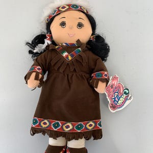 Peut inclure: Poupée faite à la main vêtue d'une robe marron en similicuir avec des bordures et des franges colorées. La poupée a des cheveux noirs en fil, un bandeau et des mocassins. Une étiquette avec un motif coloré est attachée.