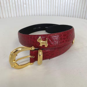 Peut inclure: Une ceinture rouge avec un motif texturé et une boucle dorée. La ceinture comporte des breloques dorées en forme de chien. La ceinture mesure 132-185 cm.