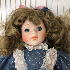 Puede incluir: Una muñeca de porcelana con cabello castaño rizado, ojos azules y un lazo rosa. Lleva un vestido azul con cuello de encaje blanco y un estampado floral rosa y azul.