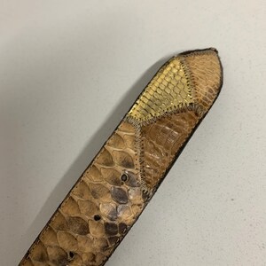 K&ouml;nnte beinhalten: Nahaufnahme eines G&uuml;rtels aus Schlangenhaut. Der G&uuml;rtel hat ein Patchwork-Design mit Braun- und Beiget&ouml;nen und einen goldfarbenen Abschnitt am Ende. Der G&uuml;rtel hat eine strukturierte Oberfl&auml;che und sichtbare N&auml;hte.