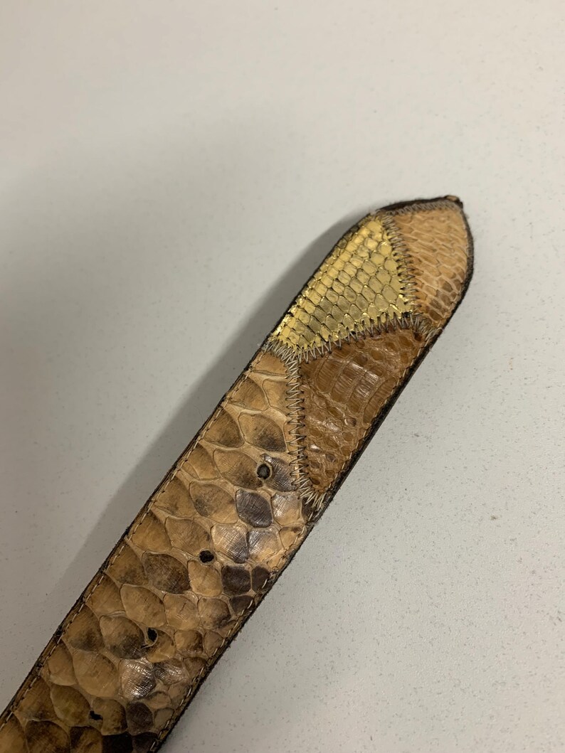 K&ouml;nnte beinhalten: Nahaufnahme eines G&uuml;rtels aus Schlangenhaut. Der G&uuml;rtel hat ein Patchwork-Design mit Braun- und Beiget&ouml;nen und einen goldfarbenen Abschnitt am Ende. Der G&uuml;rtel hat eine strukturierte Oberfl&auml;che und sichtbare N&auml;hte.