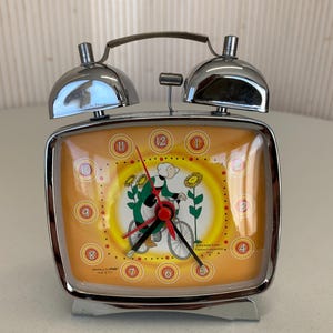 Può includere: Una sveglia vintage cromata con quadrante quadrato e due campane sulla parte superiore. Il quadrante arancione presenta un cartone animato di una donna in bicicletta. L'orologio ha lancette nere e dettagli rossi.