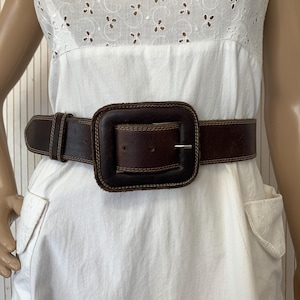 May include: A brown leather belt with a large square buckle. The belt is worn over a white dress with eyelet detailing. The buckle is a dark brown color with a gold-colored metal clasp.