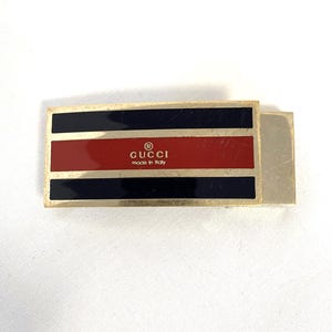 Puede incluir: Un clip de dinero rectangular Gucci en tono dorado. Presenta una franja central roja, flanqueada por franjas negras, y el logotipo de Gucci con el texto "made in Italy". Un accesorio de lujo clásico.