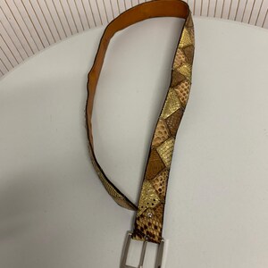 K&ouml;nnte beinhalten: Ein brauner und goldfarbener Schlangenleder-G&uuml;rtel mit einer silbernen Schnalle. Der G&uuml;rtel hat ein Patchwork-Design mit verschiedenen Braun- und Goldt&ouml;nen, wodurch ein strukturiertes Aussehen entsteht. Die Schnalle ist rechteckig und hat ein gl&auml;nzendes Finish.