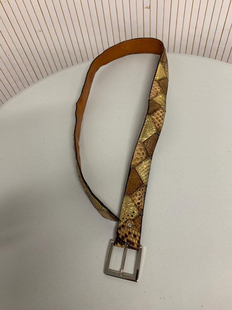 K&ouml;nnte beinhalten: Ein brauner und goldfarbener Schlangenleder-G&uuml;rtel mit einer silbernen Schnalle. Der G&uuml;rtel hat ein Patchwork-Design mit verschiedenen Braun- und Goldt&ouml;nen, wodurch ein strukturiertes Aussehen entsteht. Die Schnalle ist rechteckig und hat ein gl&auml;nzendes Finish.