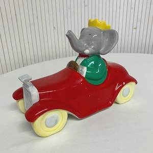 Peut inclure: Figurine en céramique d'un éléphant conduisant une voiture rouge vintage. L'éléphant est gris avec une couronne jaune, une veste verte et un nœud papillon rouge. La voiture a des roues jaunes et des accents argentés.