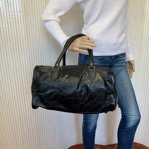 Peut inclure: Un sac de voyage en cuir noir présentant une surface texturée à panneaux avec des coutures visibles, deux poignées supérieures robustes et des détails en métal doré. Le sac a une forme rectangulaire horizontale avec des bords arrondis.