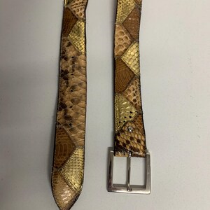 K&ouml;nnte beinhalten: Ein Patchwork-G&uuml;rtel mit silberner Schnalle. Der G&uuml;rtel besteht aus verschiedenen strukturierten Materialien in Braun-, Beige- und Goldt&ouml;nen. Das Design weist geometrische Formen und Muster auf und schafft ein einzigartiges und stilvolles Accessoire.