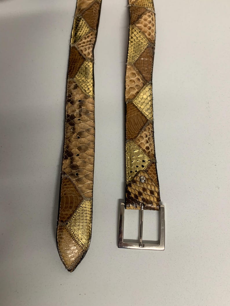 K&ouml;nnte beinhalten: Ein Patchwork-G&uuml;rtel mit silberner Schnalle. Der G&uuml;rtel besteht aus verschiedenen strukturierten Materialien in Braun-, Beige- und Goldt&ouml;nen. Das Design weist geometrische Formen und Muster auf und schafft ein einzigartiges und stilvolles Accessoire.