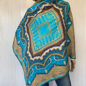 May include: A vibrant, patterned shawl in shades of turquoise, brown, and gold. The shawl features a central square design with floral motifs and intricate border details. The garment is draped over a mannequin.
