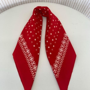 Pode incluir: Uma bandana vermelha com padrões florais e geométricos brancos. A bandana é dobrada ao meio, criando uma forma triangular. O design branco inclui pequenas flores e bordas decorativas.