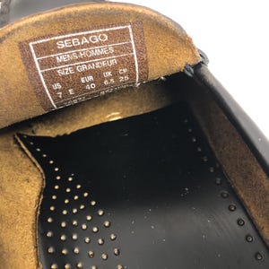 Pu&ograve; includere: Primo piano dell'interno di una scarpa in pelle nera con una soletta in pelle marrone. La soletta ha un'etichetta con la taglia che recita "Sebago Mens-Hommes Size Grandeur US EUR UK CM 7E 40 6.5 25".