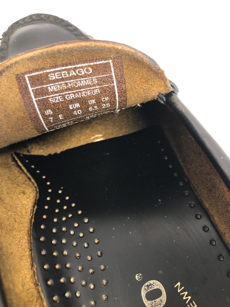 Pu&ograve; includere: Primo piano dell'interno di una scarpa in pelle nera con una soletta in pelle marrone. La soletta ha un'etichetta con la taglia che recita "Sebago Mens-Hommes Size Grandeur US EUR UK CM 7E 40 6.5 25".
