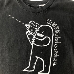 May include: Black t-shirt with a white graphic of a cartoon character holding a camera. The text "VANS Skateboarding" is written in a white circle around the character.