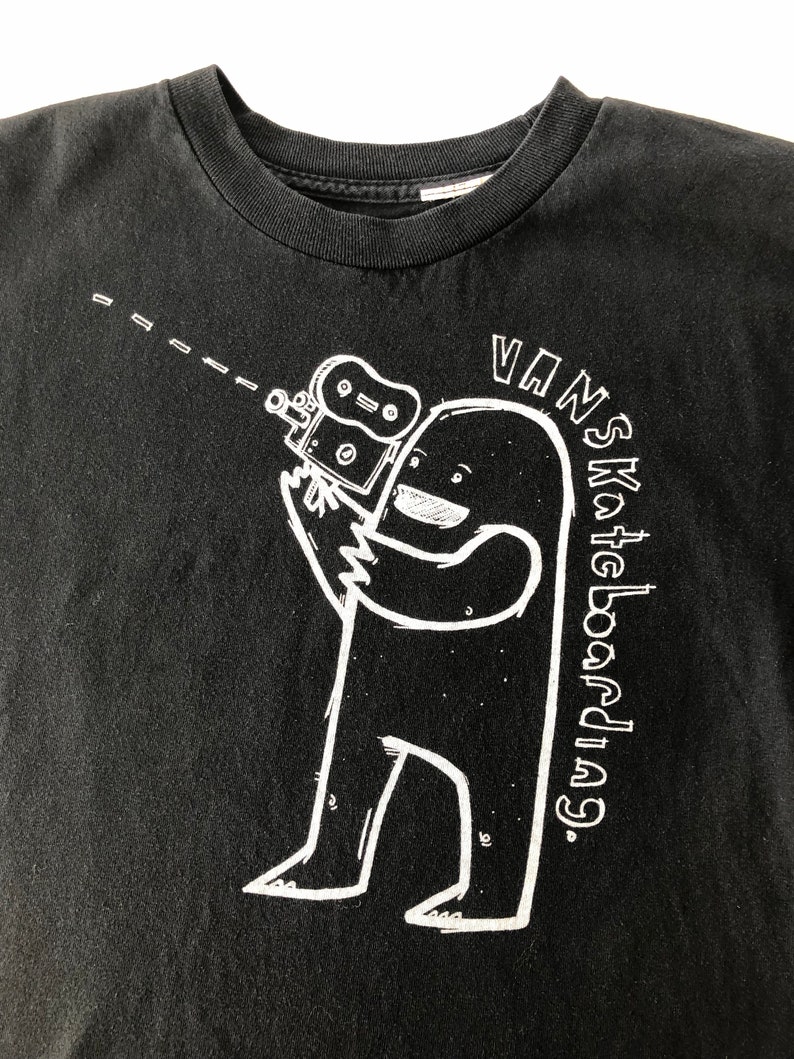 May include: Black t-shirt with a white graphic of a cartoon character holding a camera. The text "VANS Skateboarding" is written in a white circle around the character.