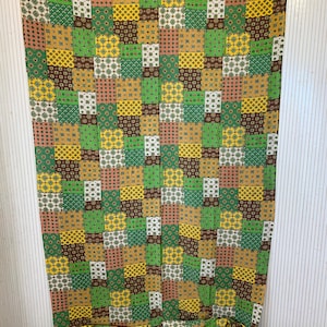 Può includere: Un tessuto patchwork con un motivo ripetuto di quadrati nei toni del verde, giallo, marrone e bianco. Il tessuto è drappeggiato, rivelando la sua consistenza e il suo design. Il tema generale è vintage o retrò.