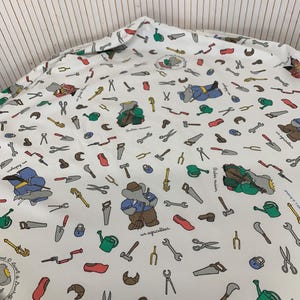 Könnte beinhalten: Weißer Stoff mit einem sich wiederholenden Muster aus Werkzeugen, Gartengeräten und Zeichentrick-Elefanten. Das Design umfasst Sägen, Scheren, Schraubenschlüssel und Gießkannen in verschiedenen Farben. Text auf Französisch ist ebenfalls sichtbar.