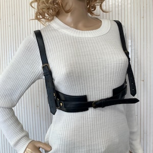 May include: Black leather harness with adjustable straps and brass-colored buckles. The harness features a chest strap and a waist belt, designed to be worn over clothing. The product is displayed on a mannequin wearing a white long-sleeved shirt.