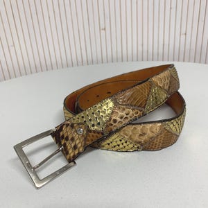 Peut inclure: Une ceinture décorative avec un motif patchwork. La ceinture présente un mélange de pièces de cuir dorées, marron et beige. Elle possède une boucle carrée de couleur argentée et une seule boucle. La ceinture est posée sur une surface blanche.