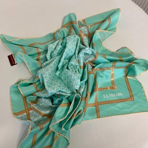 Puede incluir: Un pañuelo cuadrado plegado, en tonos turquesa y blanco, con un patrón geométrico repetido y un borde contrastante. El pañuelo presenta un diseño de correas de estilo ecuestre de color beige y el texto "U.S. POLO ASSN."