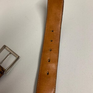 K&ouml;nnte beinhalten: Ein brauner Lederg&uuml;rtel mit einer silbernen Schnalle. Der G&uuml;rtel hat eine hellbraune Innenseite und ist mit einem helleren Faden gen&auml;ht. Die Schnalle ist rechteckig und hat eine gl&auml;nzende Oberfl&auml;che. Der G&uuml;rtel hat mehrere L&ouml;cher zur Verstellung.