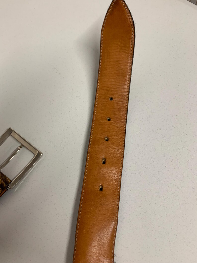 K&ouml;nnte beinhalten: Ein brauner Lederg&uuml;rtel mit einer silbernen Schnalle. Der G&uuml;rtel hat eine hellbraune Innenseite und ist mit einem helleren Faden gen&auml;ht. Die Schnalle ist rechteckig und hat eine gl&auml;nzende Oberfl&auml;che. Der G&uuml;rtel hat mehrere L&ouml;cher zur Verstellung.