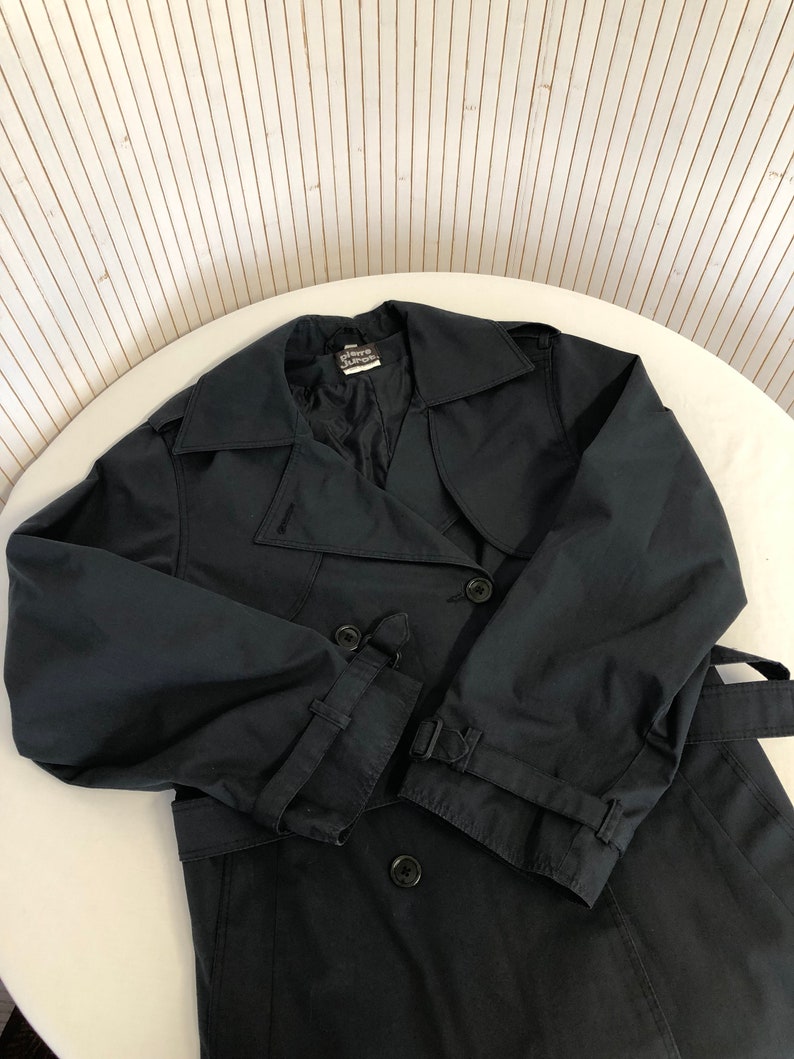 May include: A black double-breasted trench coat with a belt. The coat has a label that reads "Pierre Cardin" on the inside collar.
