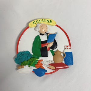 Peut inclure: Un aimant sur le thème de la cuisine avec un chef lisant un livre de cuisine. L'aimant comprend une bordure rouge et blanche, une bannière verte et jaune avec le mot "CUISINE", et des légumes, une théière et un pot.