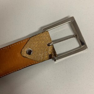 K&ouml;nnte beinhalten: Ein brauner Lederg&uuml;rtel mit einer silberfarbenen rechteckigen Schnalle. Der G&uuml;rtel hat einen Abschnitt mit Schlangenlederstruktur in der N&auml;he der Schnalle und ist mit einem Kontrastfaden gen&auml;ht.