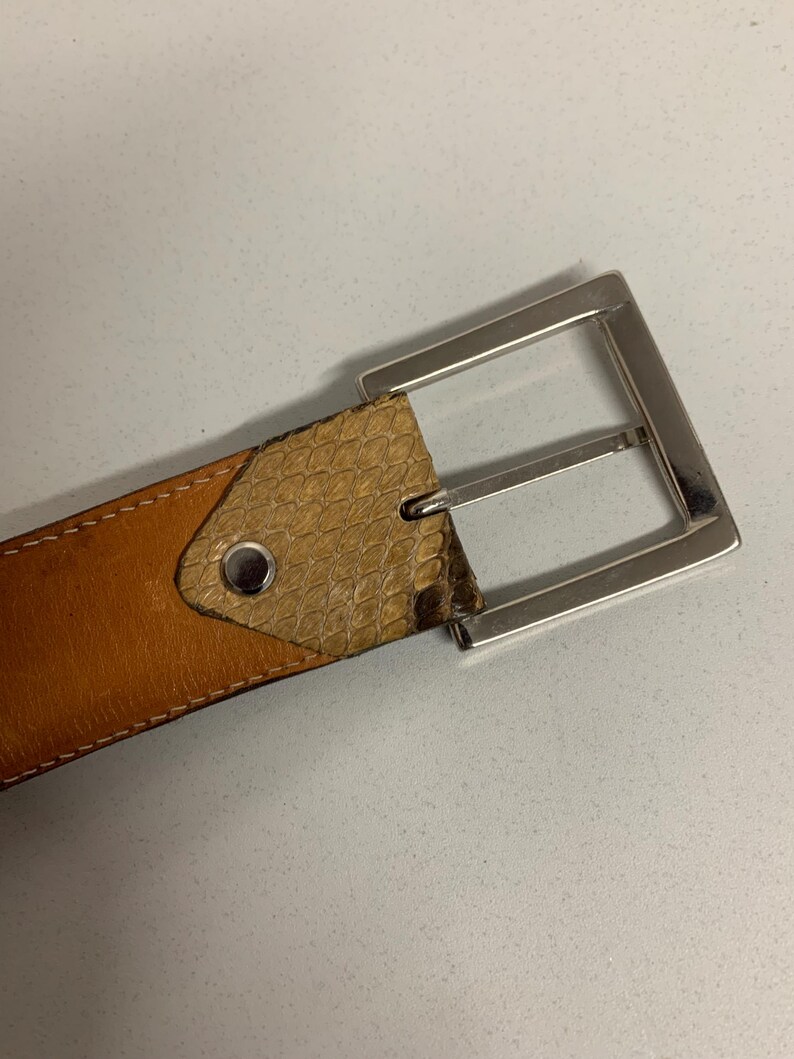 K&ouml;nnte beinhalten: Ein brauner Lederg&uuml;rtel mit einer silberfarbenen rechteckigen Schnalle. Der G&uuml;rtel hat einen Abschnitt mit Schlangenlederstruktur in der N&auml;he der Schnalle und ist mit einem Kontrastfaden gen&auml;ht.