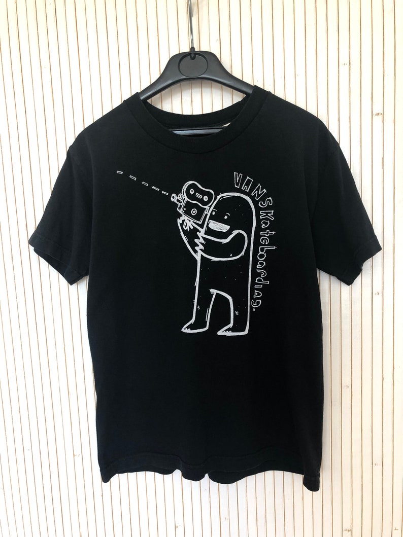 May include: Black t-shirt with a white graphic of a cartoon monster holding a camera. The text "VANS Skateboard" is written vertically on the right side of the graphic.
