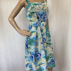 Puede incluir: Vestido hawaiano vintage con estampado tropical de palmeras y flores de hibisco azules. El vestido tiene un escote cuadrado, un cinturón blanco y una falda a la rodilla. La paleta de colores incluye azul, amarillo y verde.