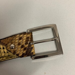 K&ouml;nnte beinhalten: Nahaufnahme eines G&uuml;rtels mit Schlangenmuster in Braun- und Beiget&ouml;nen. Der G&uuml;rtel hat eine silberfarbene rechteckige Schnalle. Der Lederg&uuml;rtel ist ein Modeaccessoire.