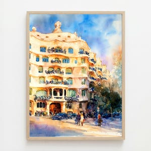 May include: Watercolor painting of a building with a unique, undulating facade. The building is a warm beige color with blue windows and balconies. The sky is a mix of blue and orange, and there are people and cars on the street below. The artwork is framed in a light wood frame.