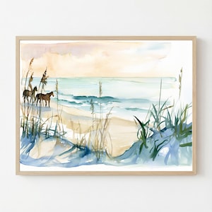 May include: A framed watercolor painting depicting a coastal beach scene with two brown horses standing on the sand. The artwork features tall green beach grass in the foreground, a light blue ocean with gentle waves, and a soft peach-colored sky.