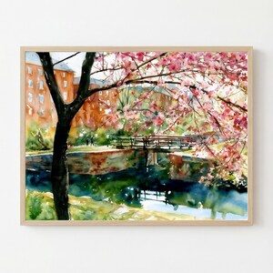 May include: A watercolor painting in a light wood frame depicting a canal scene with a dark tree trunk in the foreground, pink cherry blossoms, a stone bridge, and orange brick buildings reflected in the dark blue water under a bright sky.