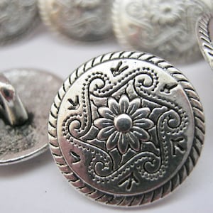 May include: Silver metal button with a decorative floral design. The button has a raised edge and a smooth, shiny finish.