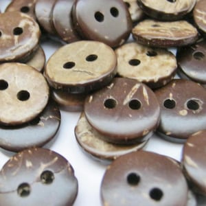 以下が含まれることがあります： 2つの穴が空いた茶色のココナッツの殻のボタンのコレクション。ボタンは白い表面にランダムなパターンで配置されています。