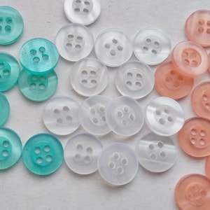 May include: A collection of assorted buttons in three colours: light blue, white, and light pink. The buttons are all round and have four holes.