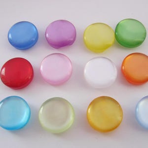 May include: Assortment of 12 round, flat, plastic buttons in various colors, including blue, purple, yellow, green, red, pink, white, orange, light blue, light green, light yellow, and gold.