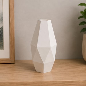 Könnte beinhalten: Eine matte weiße Vase mit einer geometrischen, facettierten Oberfläche, einem verjüngten Hals und einem breiten, kantigen Körper. Das Objekt hat eine solide, undurchsichtige Konstruktion mit scharfen, klaren Kanten, die verschiedene Dreiecke bilden.