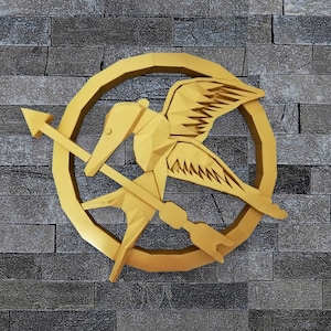 May include: A gold-colored, three-dimensional mockingjay pin with a stylized arrow piercing through the bird's body. The pin is set against a gray brick wall.