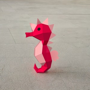 May include: A pink and red origami seahorse with a black eye. The seahorse is made of paper and has a 3D design.