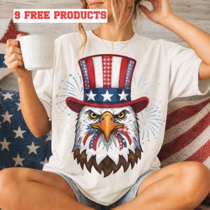 Op de afbeelding: Crèmekleurig T-shirt met een patriottisch adelaarsontwerp. De adelaar draagt een rood, wit en blauwe hoge hoed en heeft een rood, wit en blauw lint om zijn snavel. Het shirt heeft bovenaan de tekst "9 FREE PRODUCTS".
