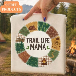 May include: A cream-colored blanket with a circular design featuring a tent, trees, mountains, and a map. The text "TRAIL LIFE MAMA" is centered within the design. The blanket is held up in front of a blurred outdoor background.