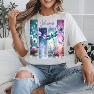 Puede incluir: Camiseta blanca con un diseño gráfico colorido de libélulas, flores y el texto "Just wing it". El diseño incluye paneles verticales con efecto acuarela, mostrando una escena serena inspirada en la naturaleza. La camiseta está siendo usada.