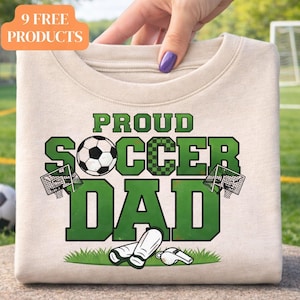 Puede incluir: Una camiseta beige con las palabras "PROUD SOCCER DAD" en letras verdes. El diseño incluye un balón de fútbol, porterías de fútbol, espinilleras y un silbato. La camiseta está sujeta por una mano con esmalte de uñas morado.